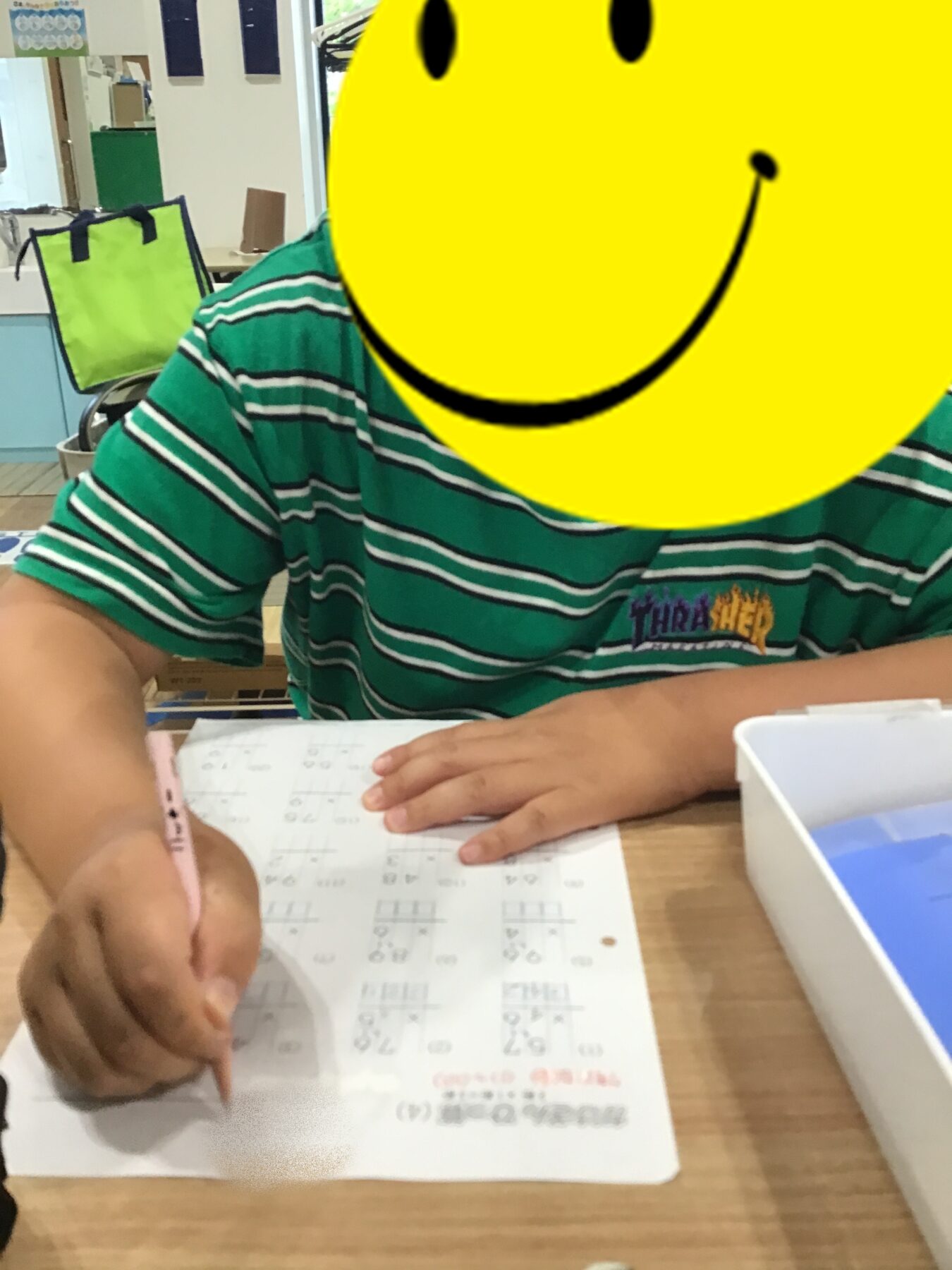 Child in a green striped shirt writes with a pink pencil at a desk, working on a math worksheet; a bright yellow smiley overlay covers their face in a classroom setting with a lime-green bag in the background.