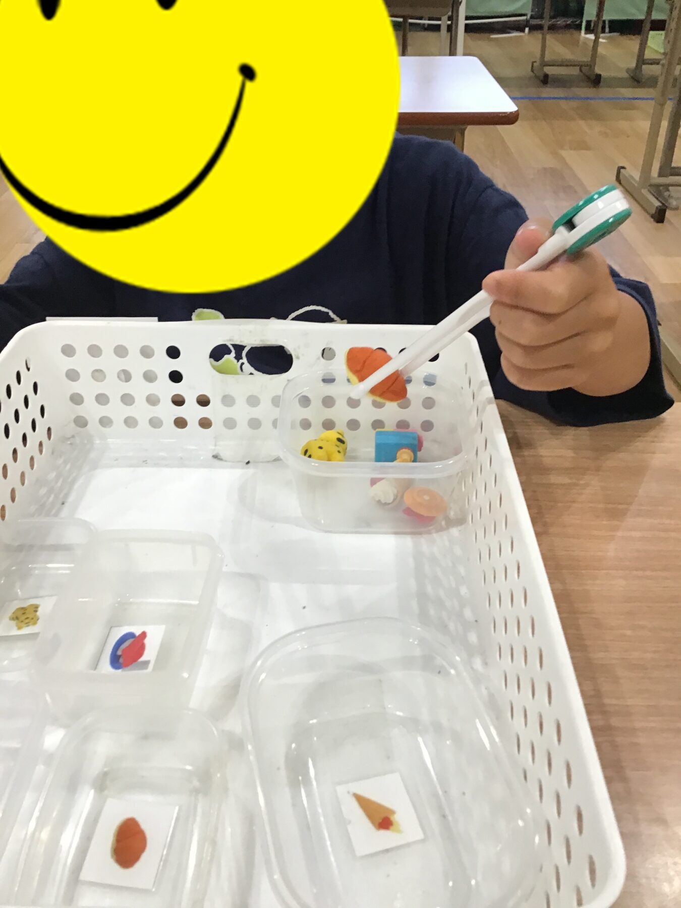 Child in a classroom uses green-and-white tongs to pick up small toy foods from a white perforated basket on a table.
