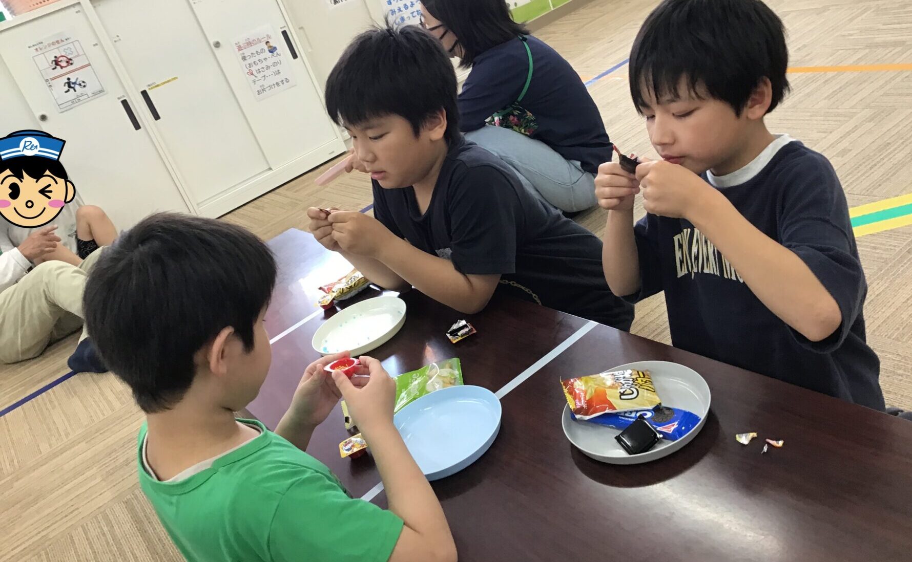 Children sit around a low table eating snacks from wrappers in a bright room with lockers along the wall in the background.