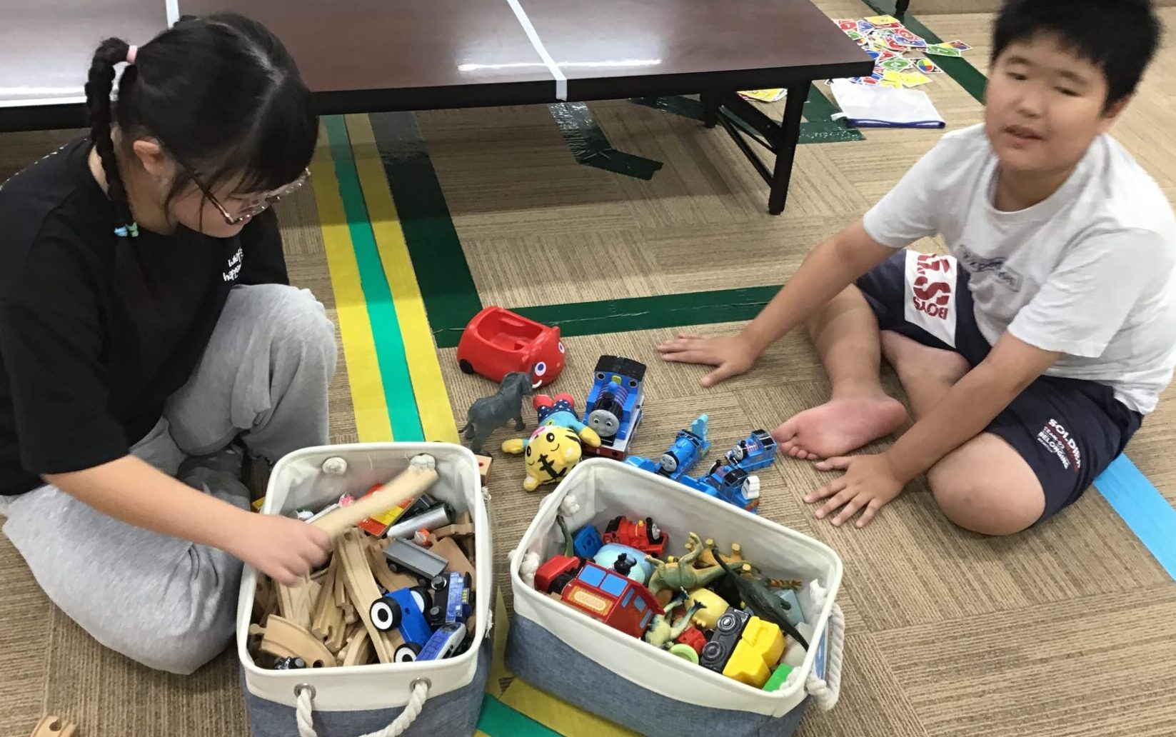 Two kids sit on the floor playing with toy trains and wooden blocks from two storage bins inside a room with a table nearby.