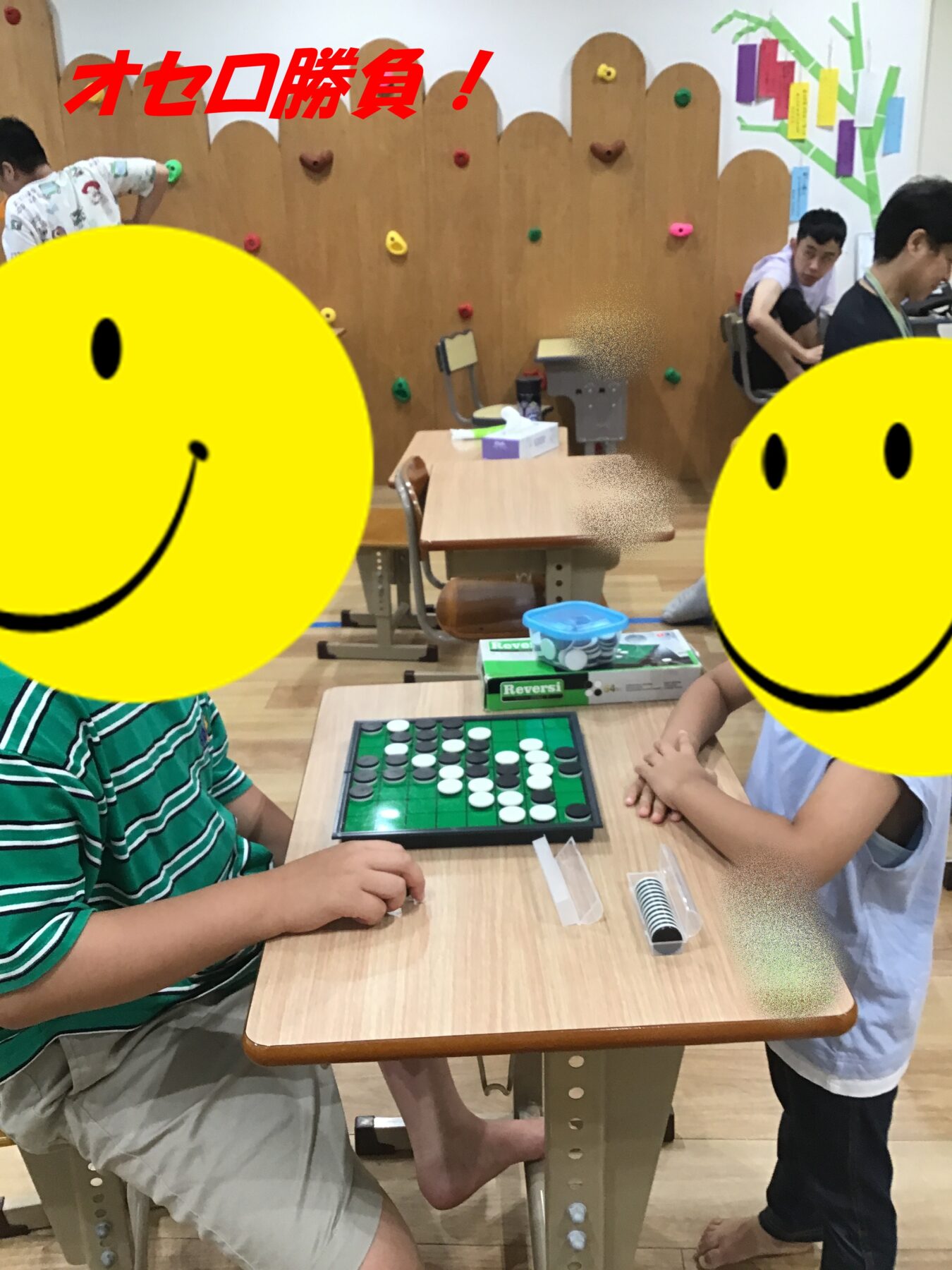 Two children sit at a wooden desk playing a game of Othello in a classroom; others watch in the background.