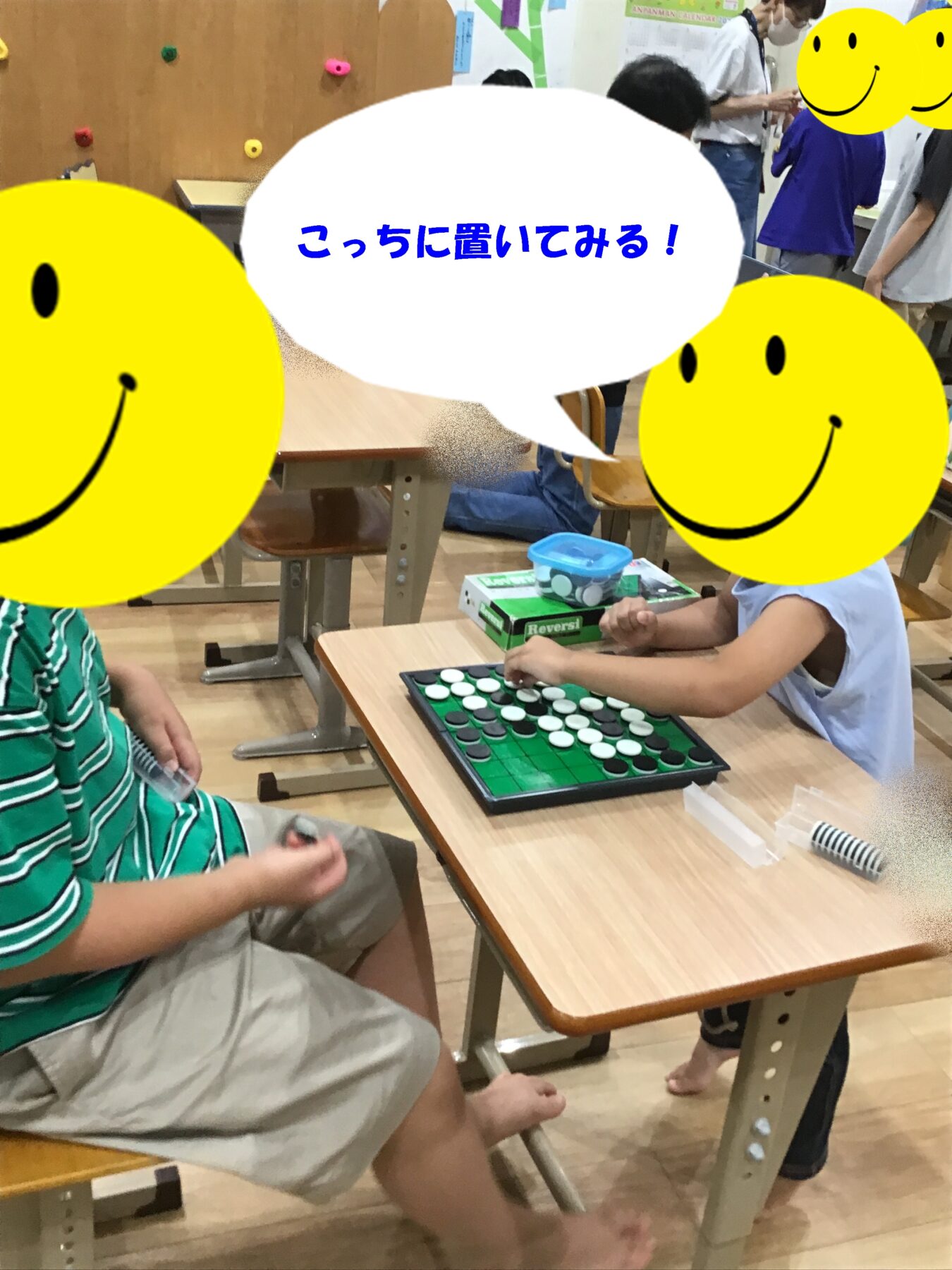 Children seated in a classroom playing an Othello game at a desk; game pieces visible on green board, with others in background and smiley stickers covering faces.