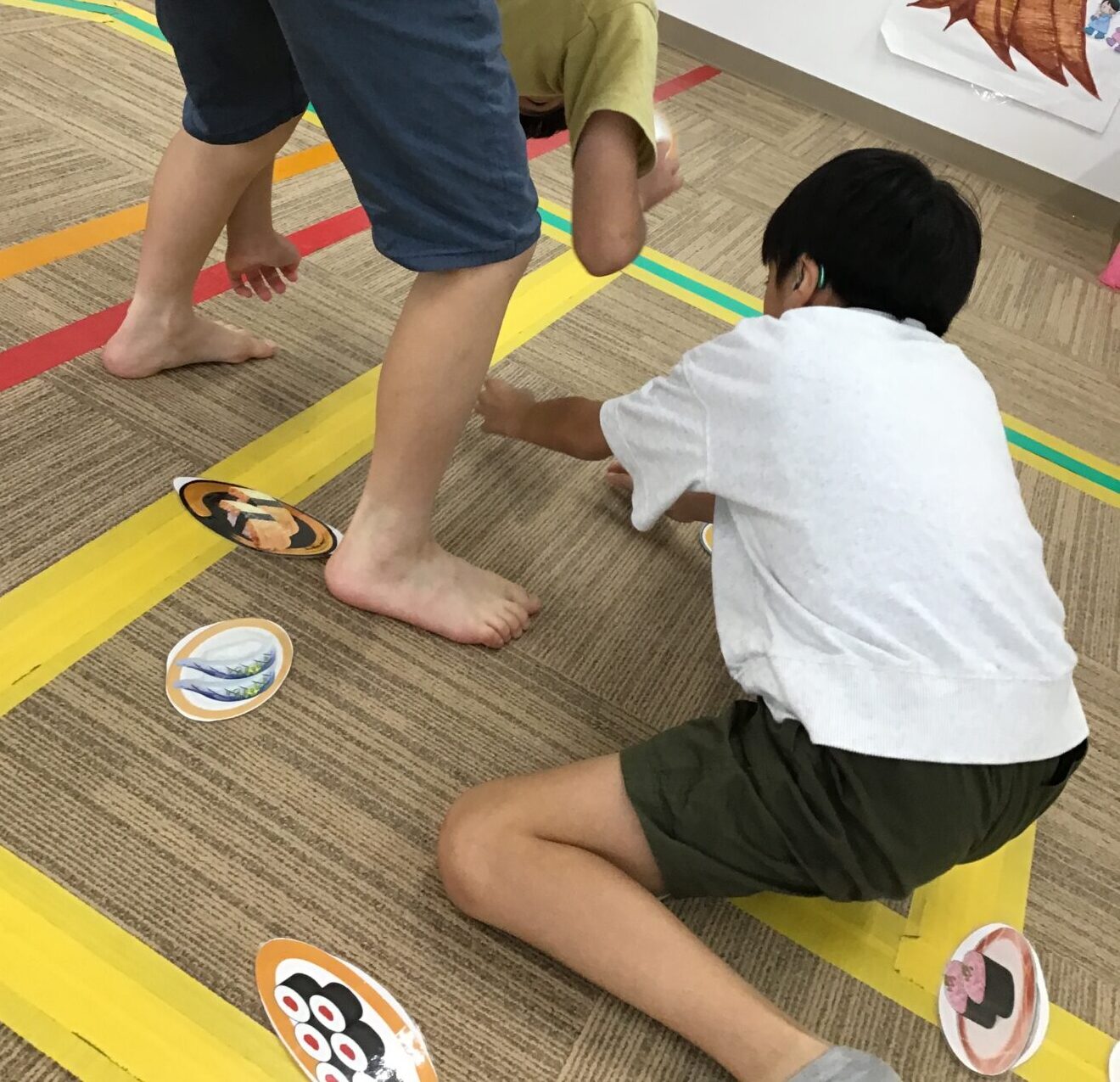 Two kids bend over on a floor game, placing or picking round cards along a yellow tape path in a classroom.