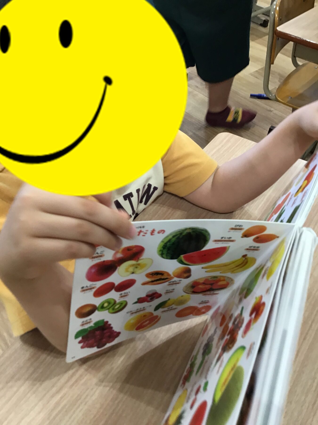 Child in a classroom looks at a colorful fruit poster/book showing various fruits on a wooden desk.