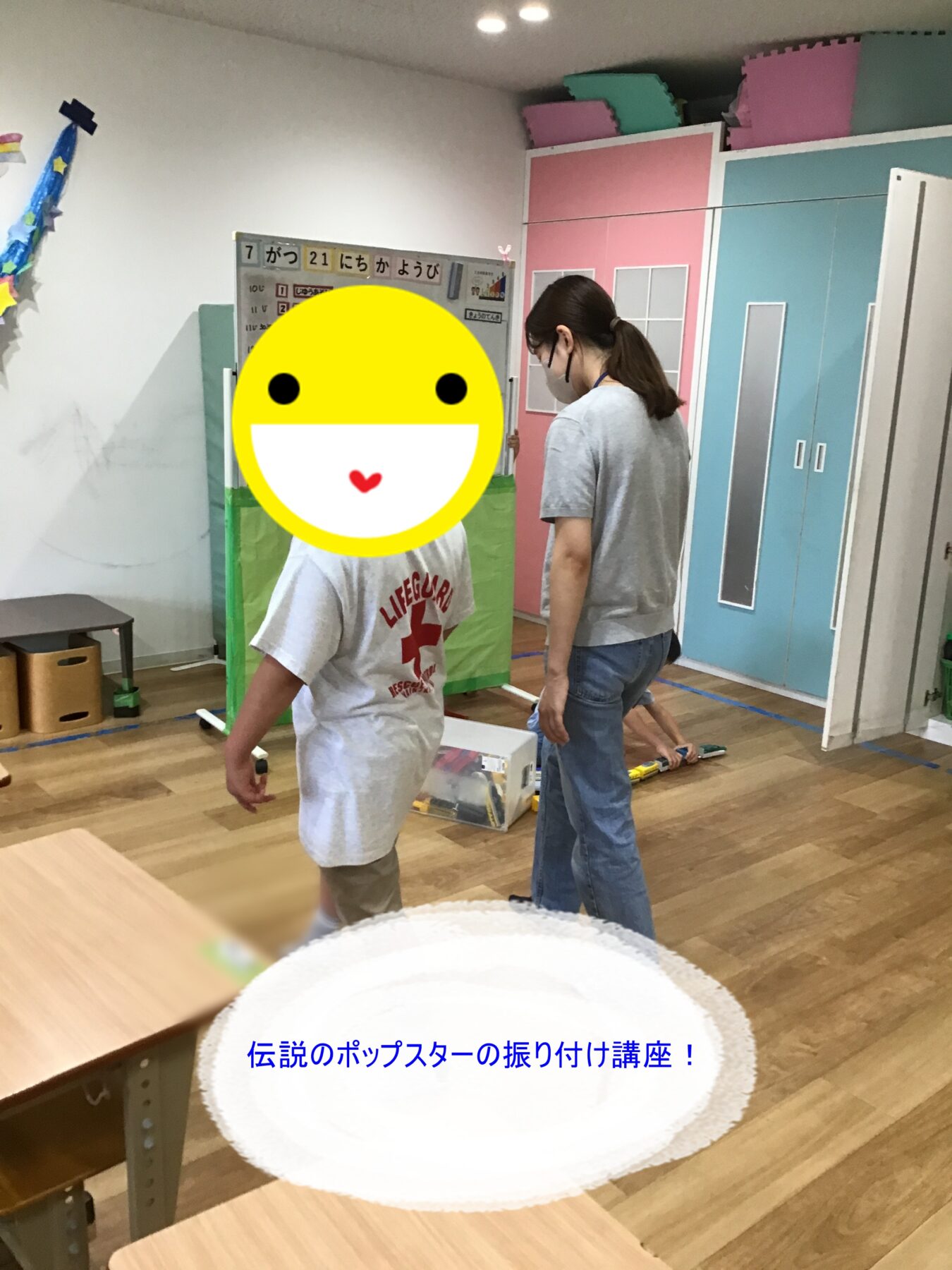 Child in a gray shirt moves in a colorful room during a dance class, with a large yellow smiley sticker over their face and a supervising adult nearby; bannered Japanese text visible in background.