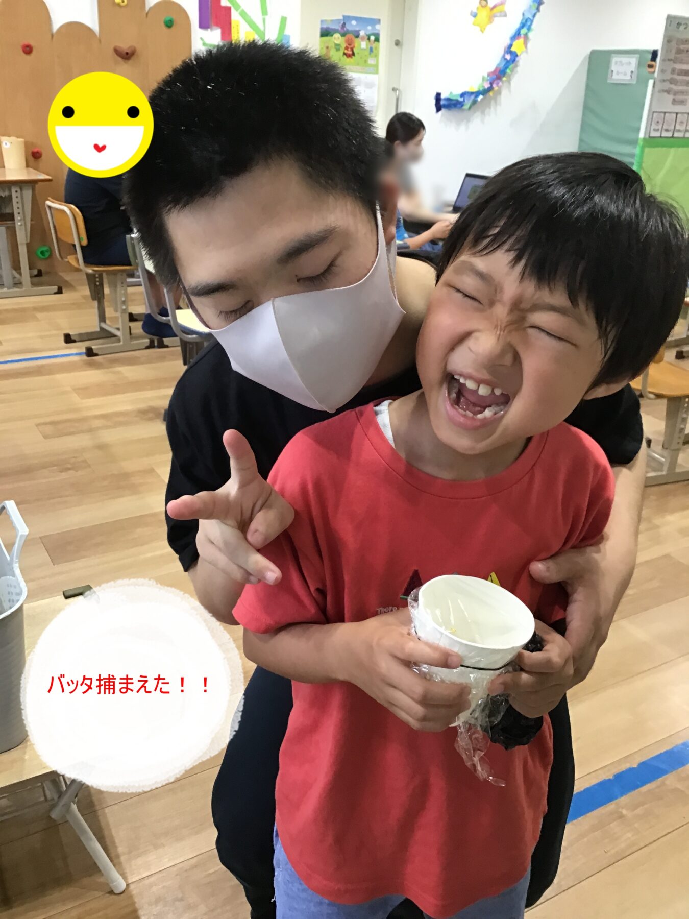 Adult caregiver wearing a white mask hugs a laughing young boy in a red shirt, as they hold a white cup together in a classroom celebration (grasshopper caption visible in red).