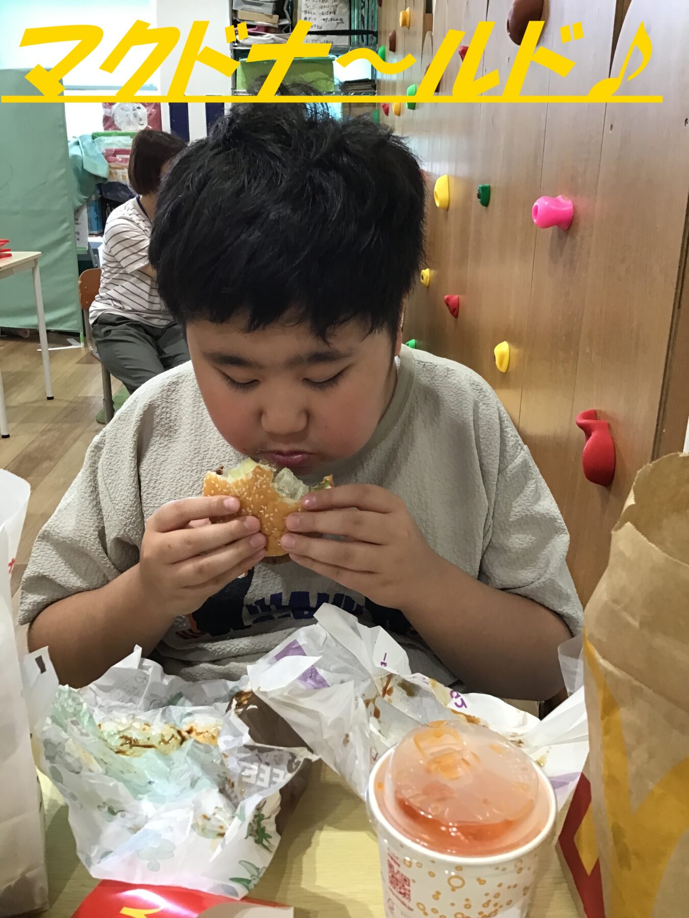 Young boy with black hair eating a burger at a fast-food table, surrounded by wrappers and a drink cup in front.