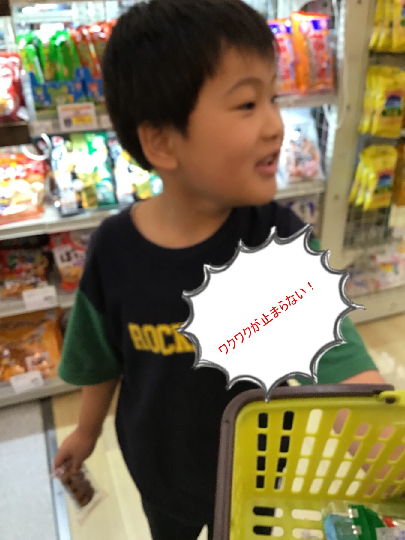 Young boy in a store, looking to the side, shelves stocked with snacks in the background, white comic-style speech bubble says excitement in Japanese.