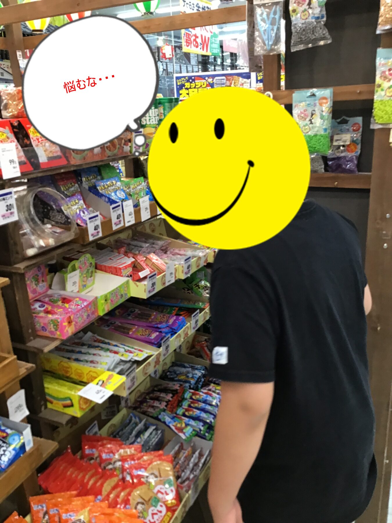 Person in a black shirt stands in a candy shop aisle browsing colorful packaged sweets on wooden shelves; a large yellow smiley graphic covers their face.