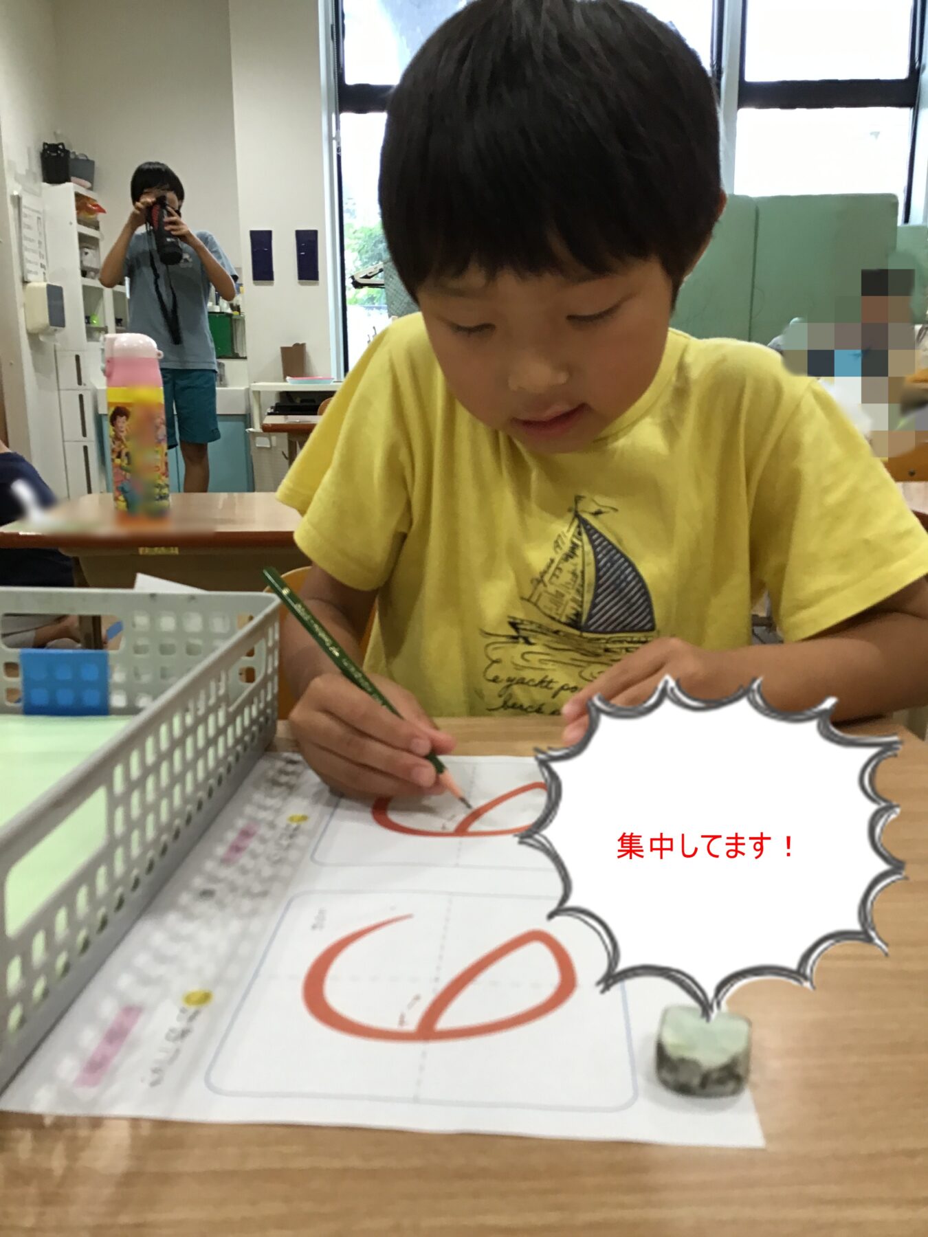 Young child in a yellow shirt focused on drawing with a green pencil at a desk in a classroom; a person in blue is taking a photo in the background.