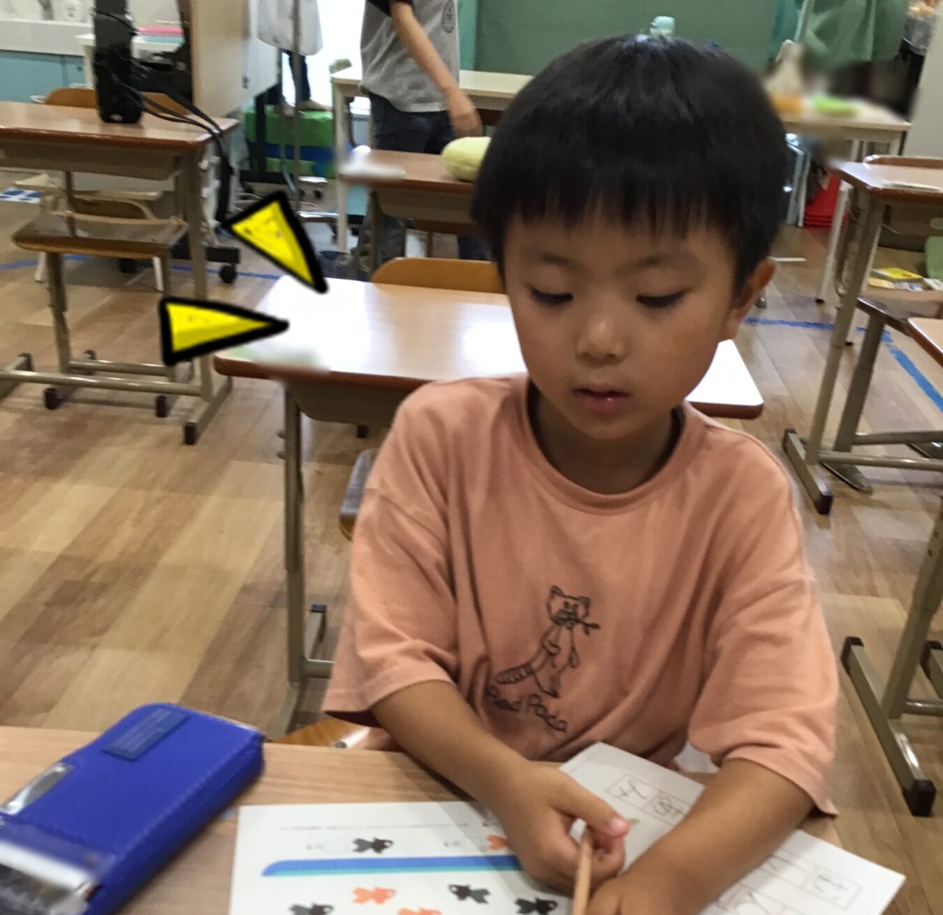 Young boy sits at a desk in a classroom, tracing shapes on a worksheet with a pencil and blue pencil case nearby.