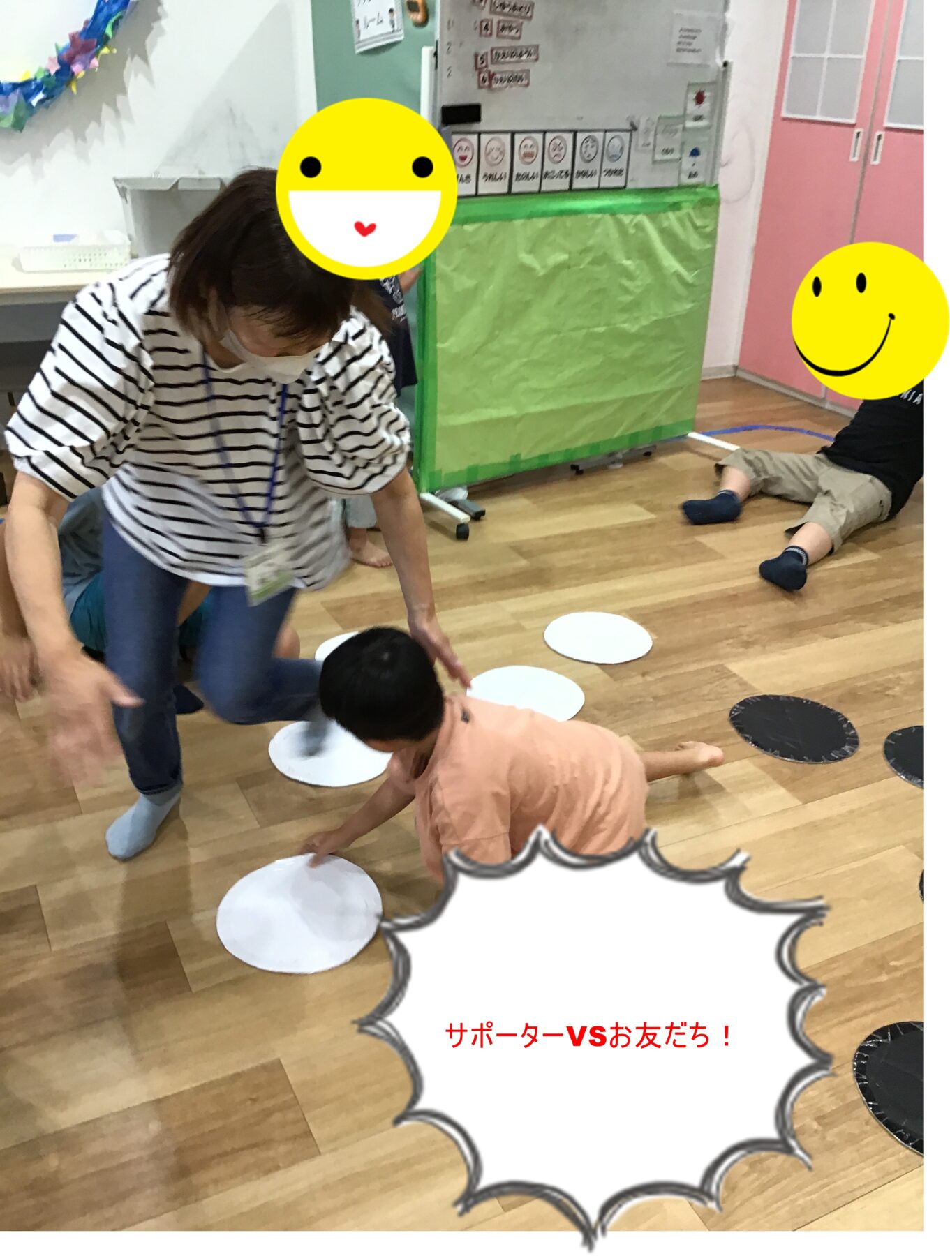 Teacher helping a child on a wooden-floored room with white paper circles on the floor during a group activity in a classroom. A second child sits nearby against a pink cabinet. Smiley stickers cover faces for privacy.