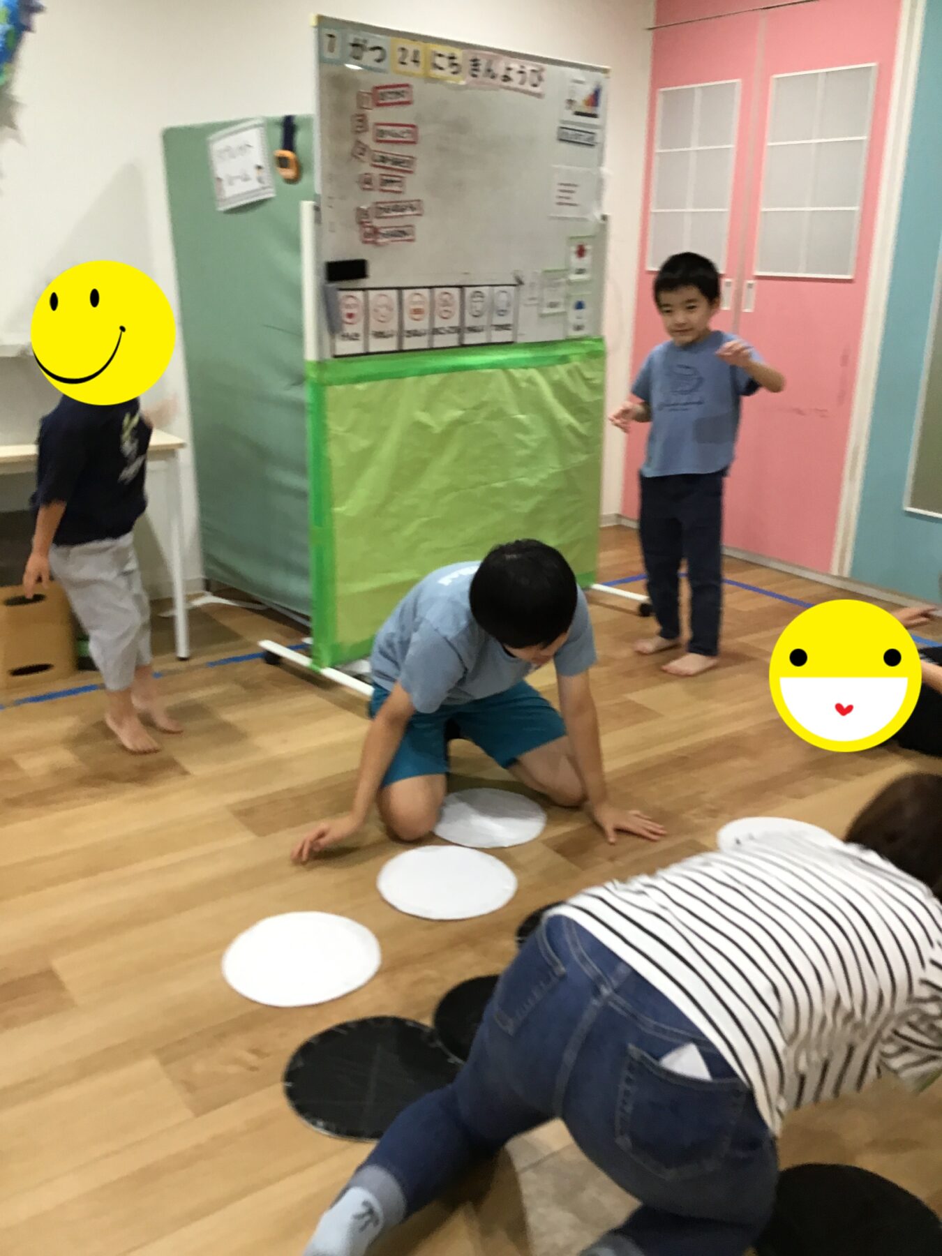Children in a classroom placing white circular mats on the wooden floor, with a bulletin board and padded wall behind them.