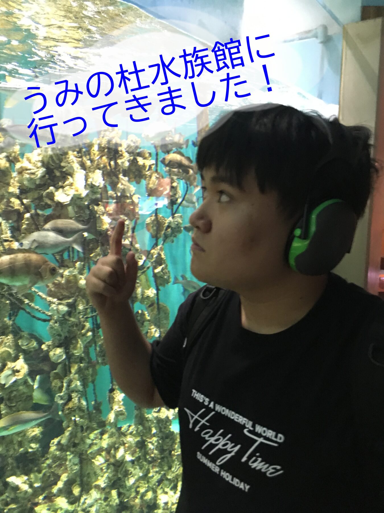 Young man wearing green headphones giving a thumbs up in front of a large aquarium filled with fish with a blue overlay text in Japanese.