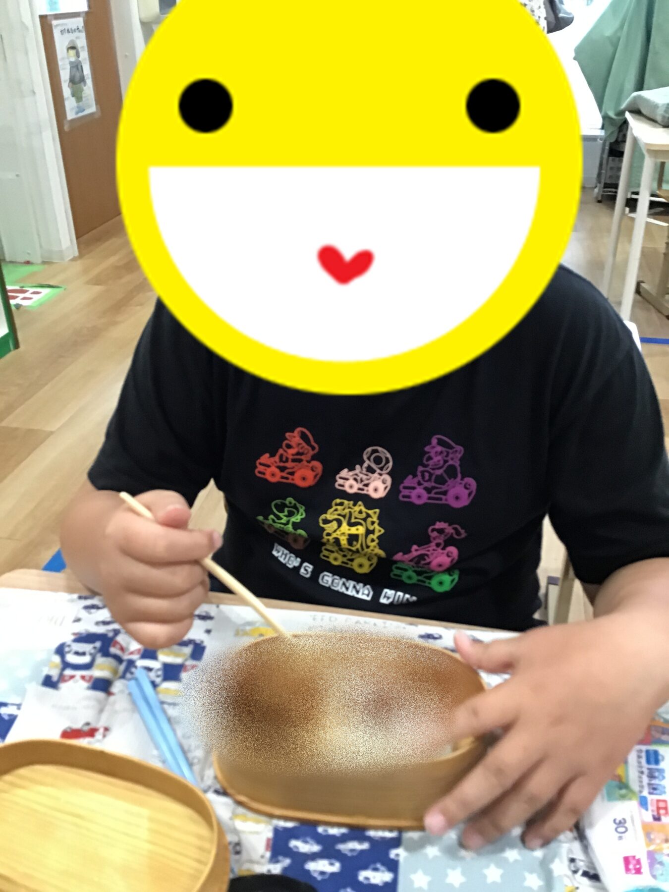 Person with a large yellow smiley overlay eating soup with chopsticks at a table indoors, wearing a black t-shirt with colorful racing car graphics.