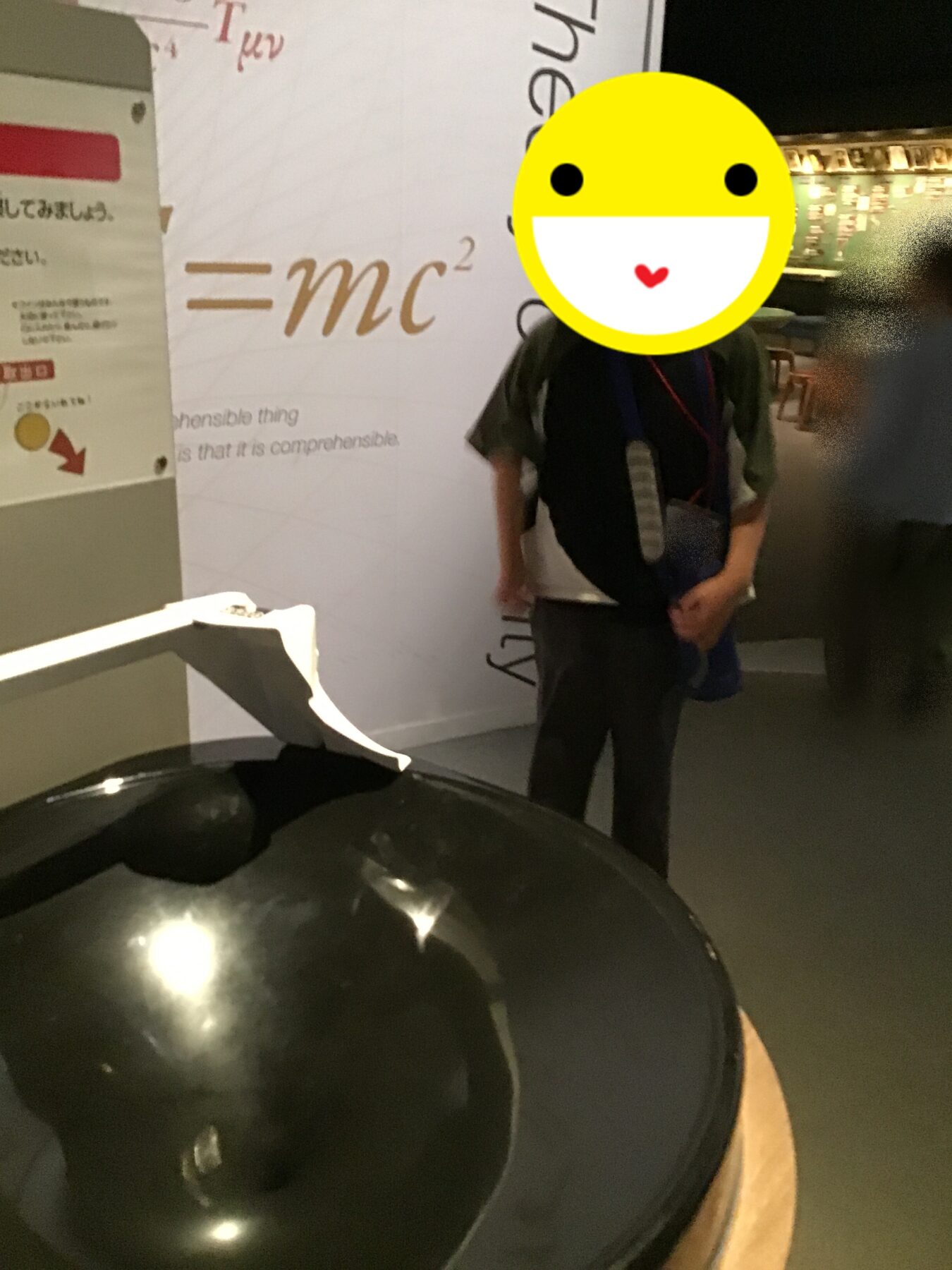 Person standing beside a wall with the equation E=mc²; a large yellow smiley mask covers their face, with a shiny black sphere in the foreground.