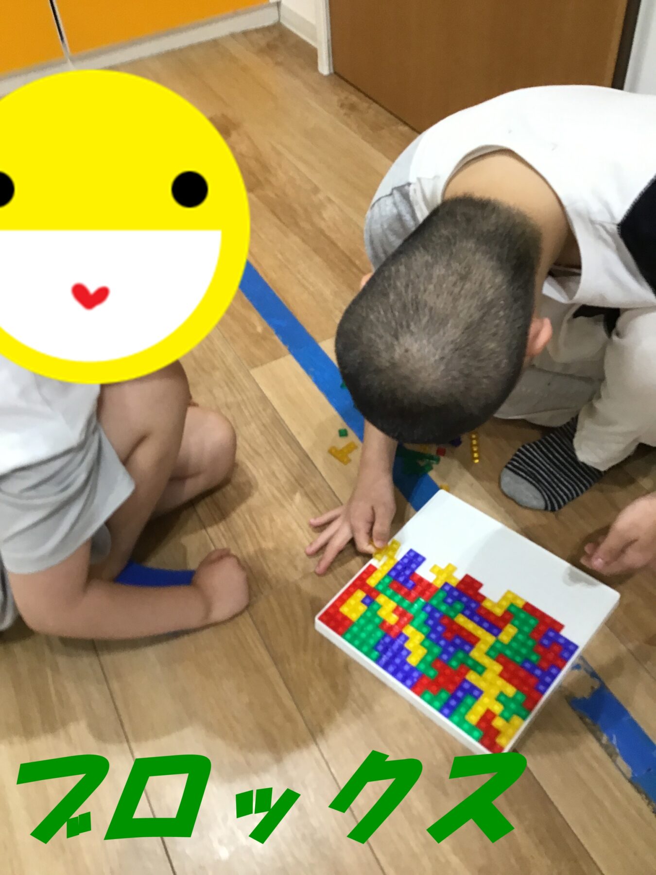 Two children sit on a wooden floor assembling colorful interlocking blocks; a large yellow smiley sticker covers the face of the child on the left.
