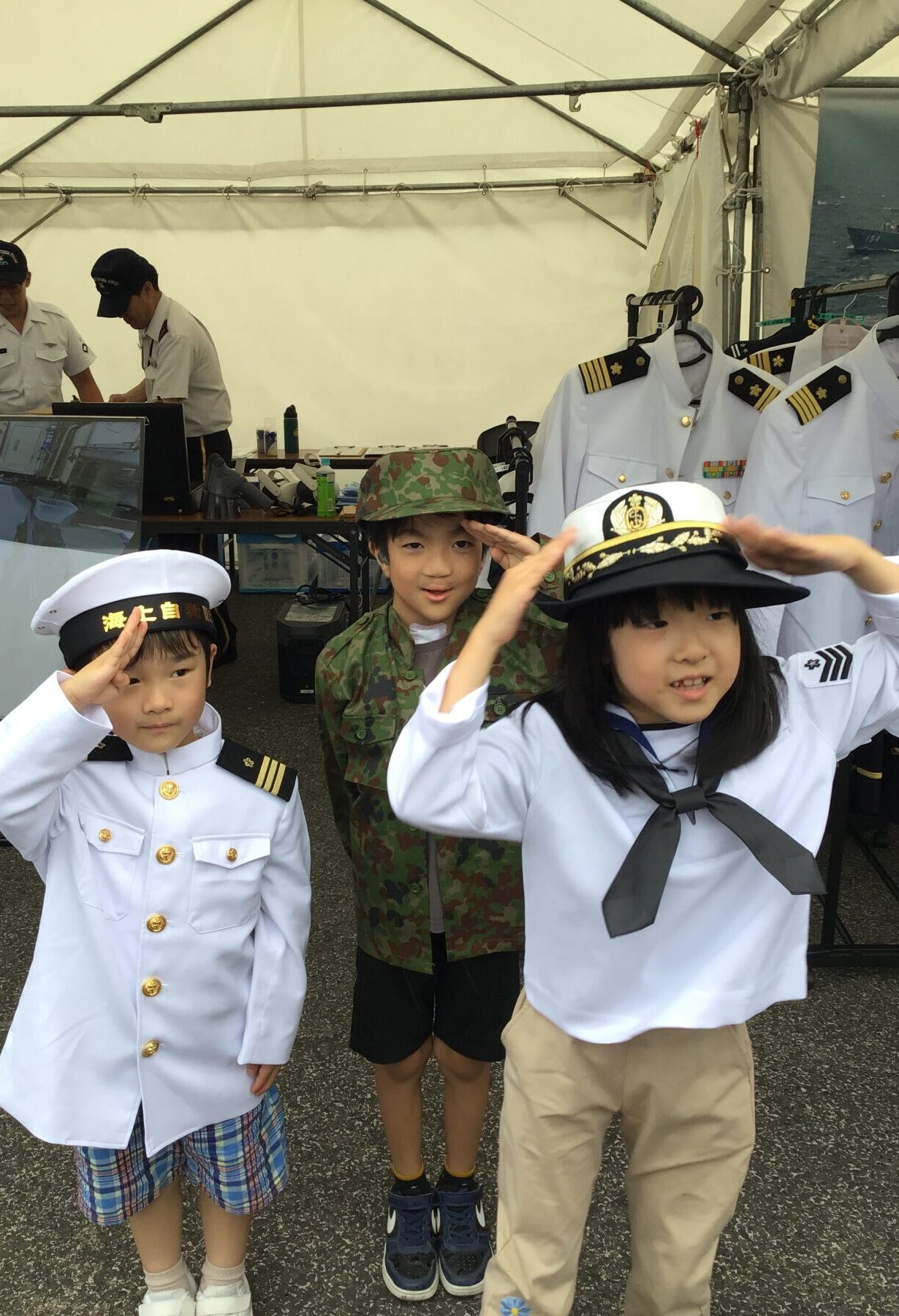 Three children in naval-style uniforms salute at the camera; a camouflage-clad child stands between them, with adults and a clothing display in a white tent behind.