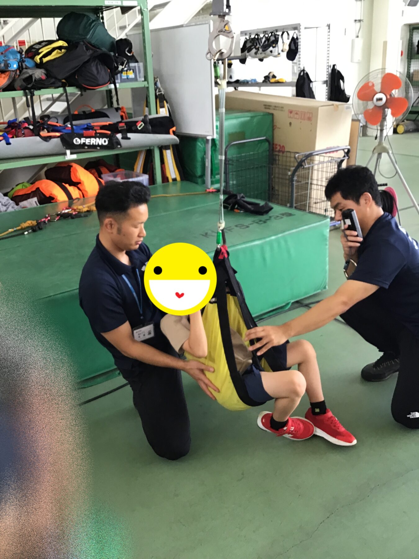 Two instructors guide a trainee in a harness during indoor safety training in a gear-filled gym setting in the background.