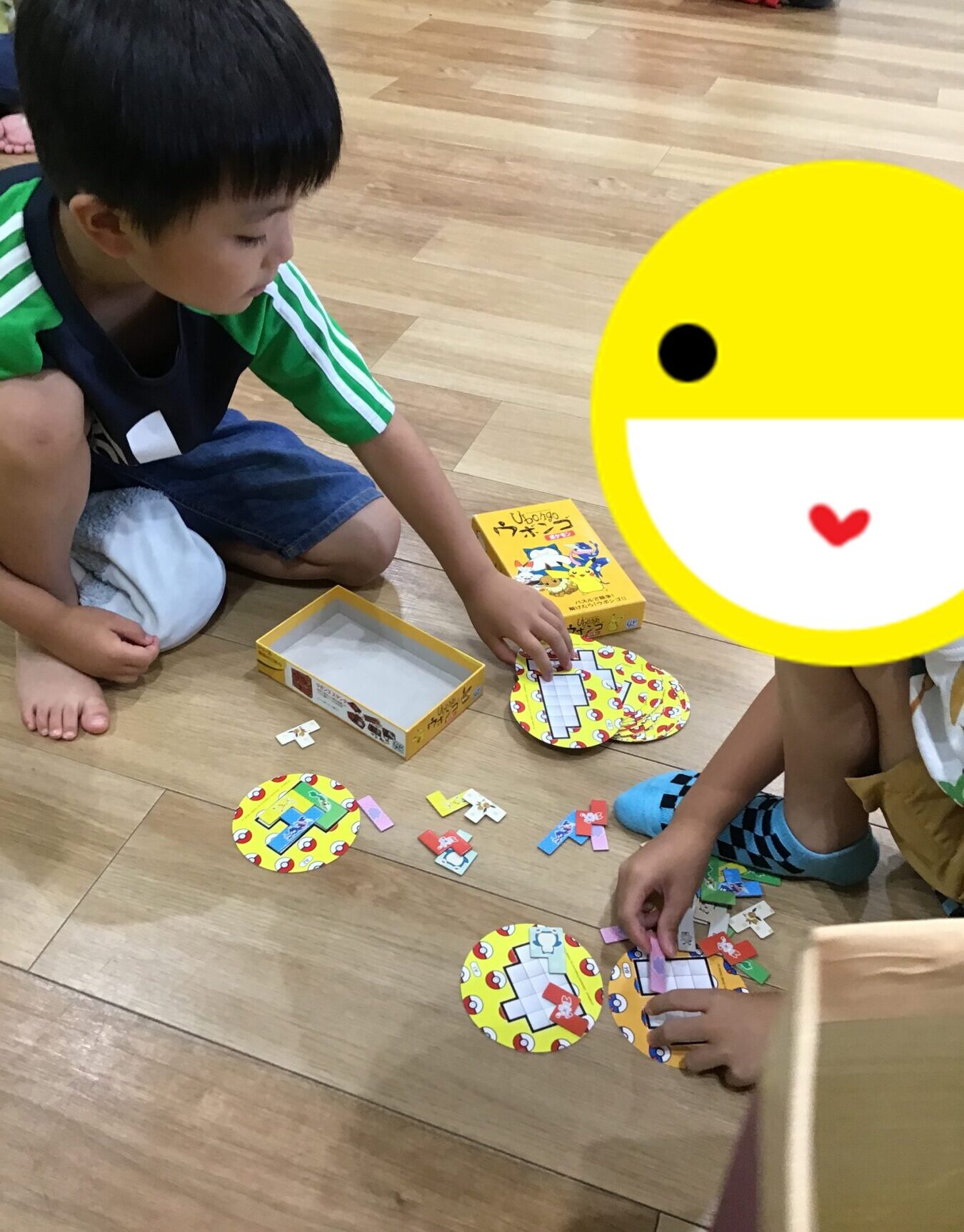 Two kids sit on a wooden floor, sorting colorful circular game cards and small pieces from a yellow box.