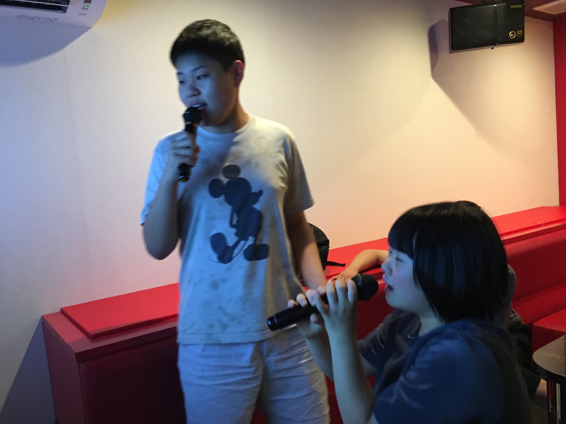 Two youths singing karaoke: one stands with a microphone wearing a light gray Mickey Mouse T‑shirt, the other sits nearby with a microphone beside red seating.