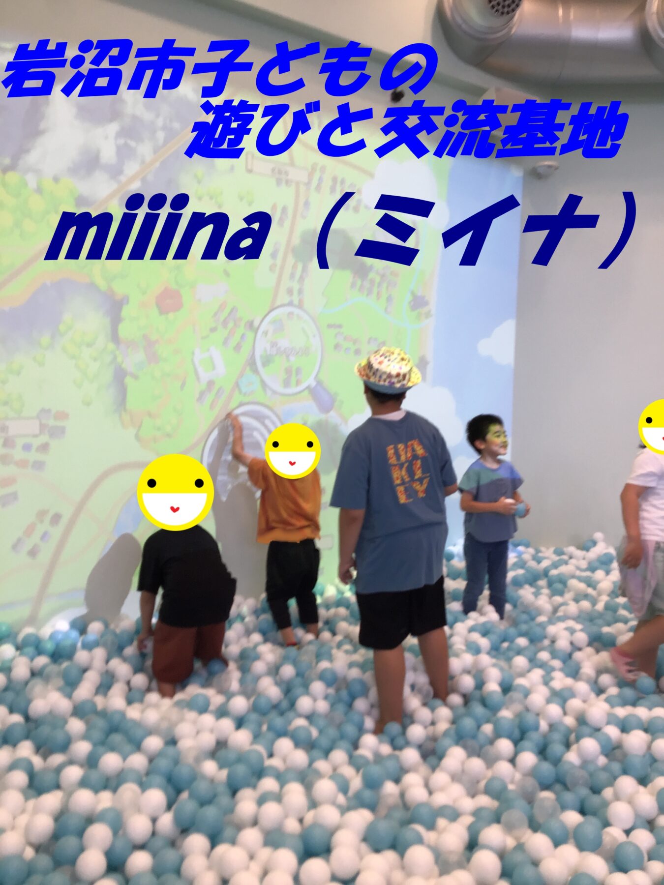 Children playing in a ball pit filled with blue and white balls inside an indoor play area; a mural with large blue Japanese text dominates the background, and some faces are covered with smiley stickers.