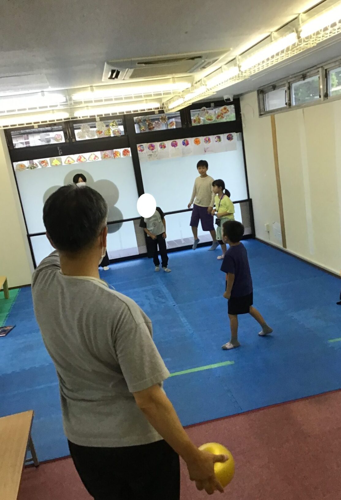 Back view of an adult holding a yellow ball, watching kids play dodgeball in a blue-matted gym floor.