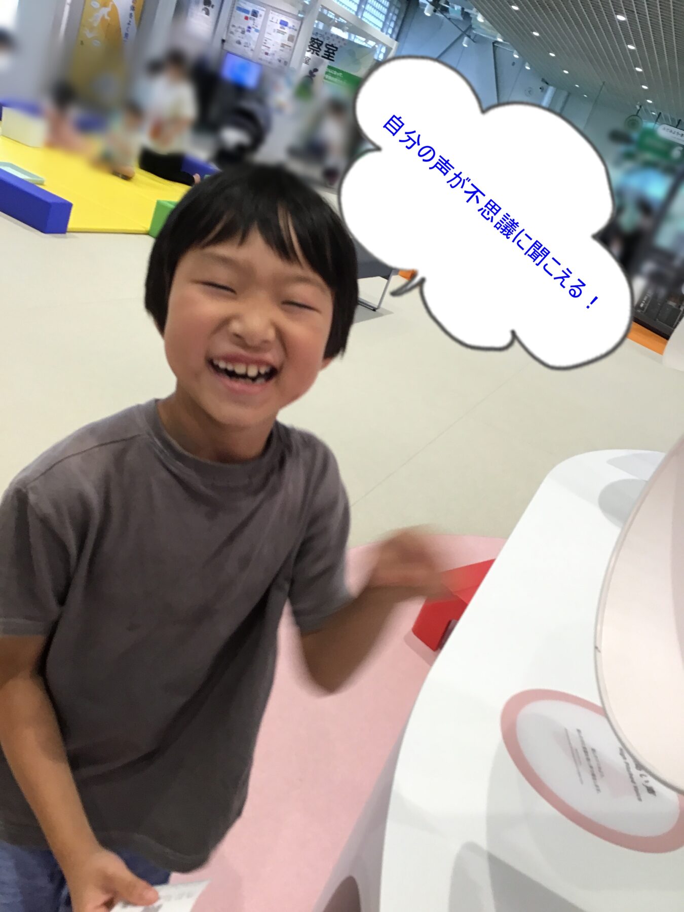 Joyful boy in a gray shirt laughing at the camera in a bright indoor play area with colorful foam blocks in the background.