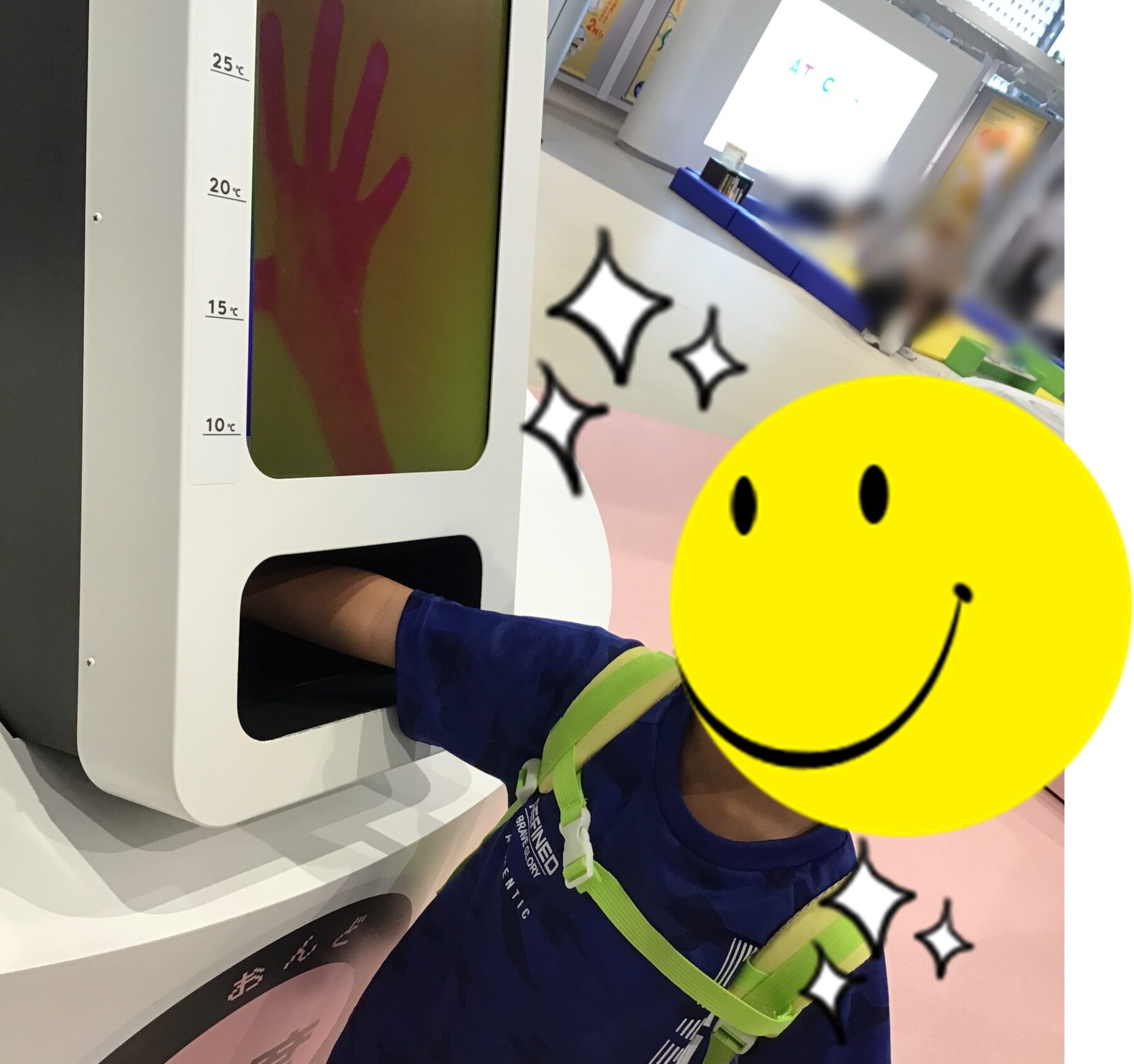 Child inserts hand into a white temperature screening device with a vertical 10–25°C scale; blurred background.