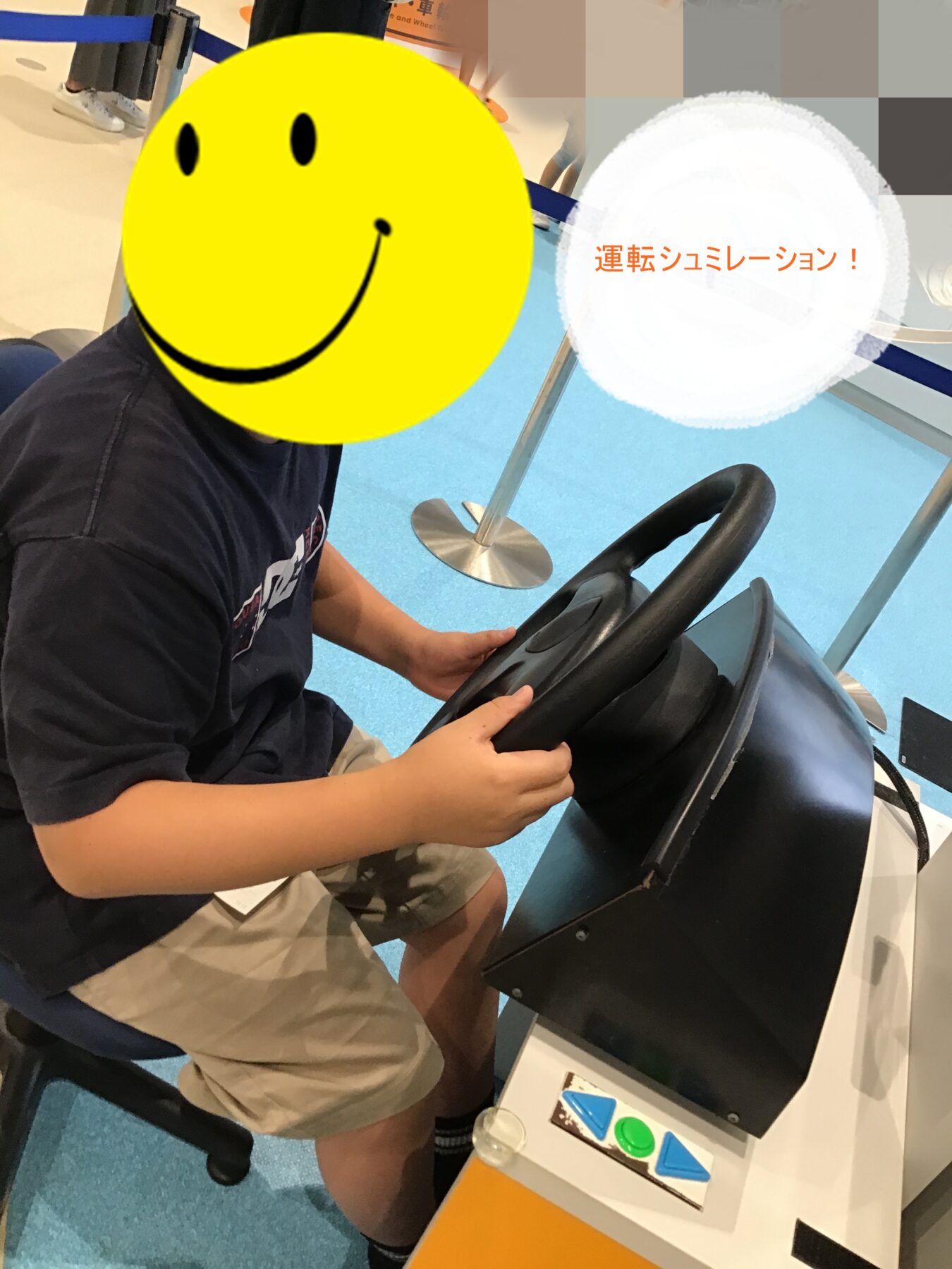 Person seated at a driving simulator holding a steering wheel with a large yellow smiley face overlay; Japanese text reads '運転シミュレーション!' in a white circle.
