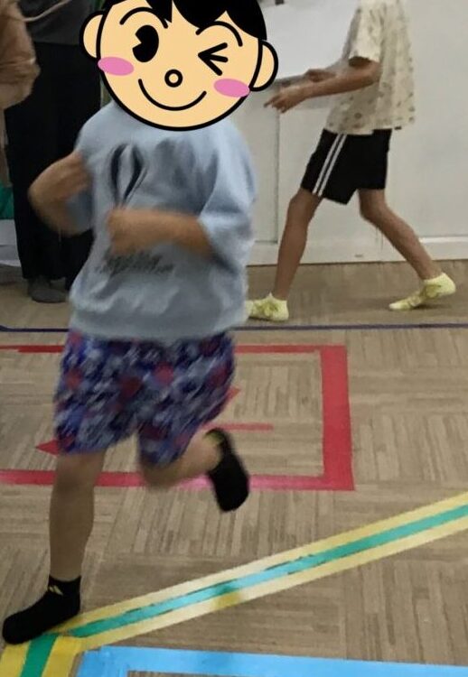 Children running along a color‑coded indoor floor track with footprints, as adults supervise in the background in a hallway.