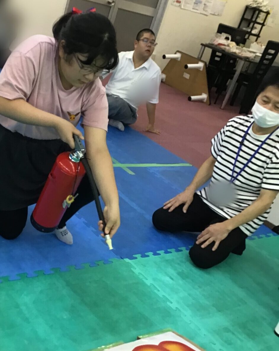 Girl kneels on blue mats, aiming a small fire extinguisher at a fire mock card on the floor during a safety drill in a classroom gym.