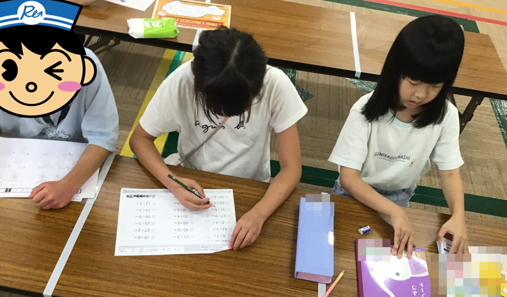 Three children sit at a wooden table completing worksheets; one has a cartoon face sticker over their face, with pencils and a purple workbook on the desk.