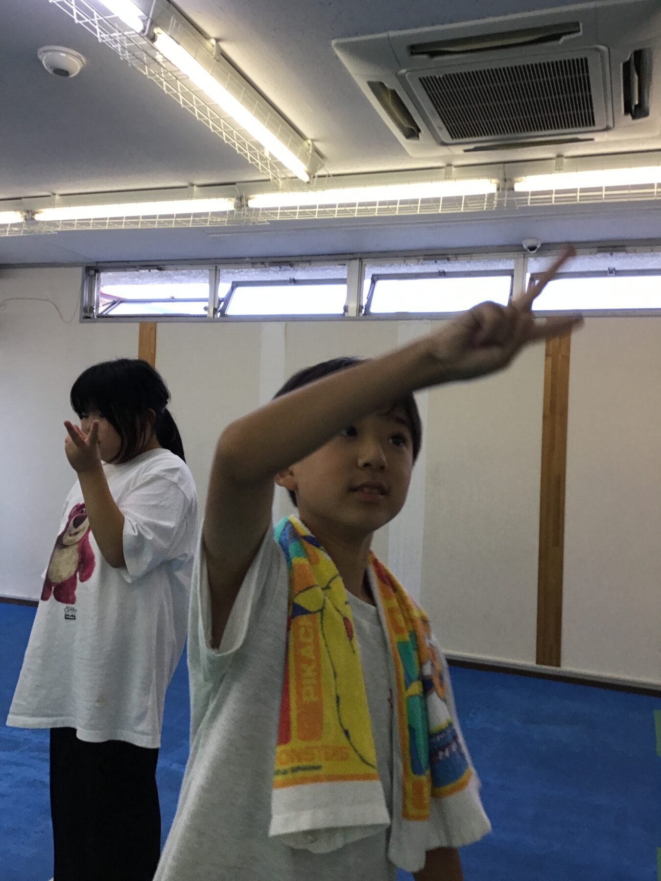 Two kids in white T-shirts stand on a blue gym mat, the foreground boy raises an arm toward the camera while a towel is draped around his neck.