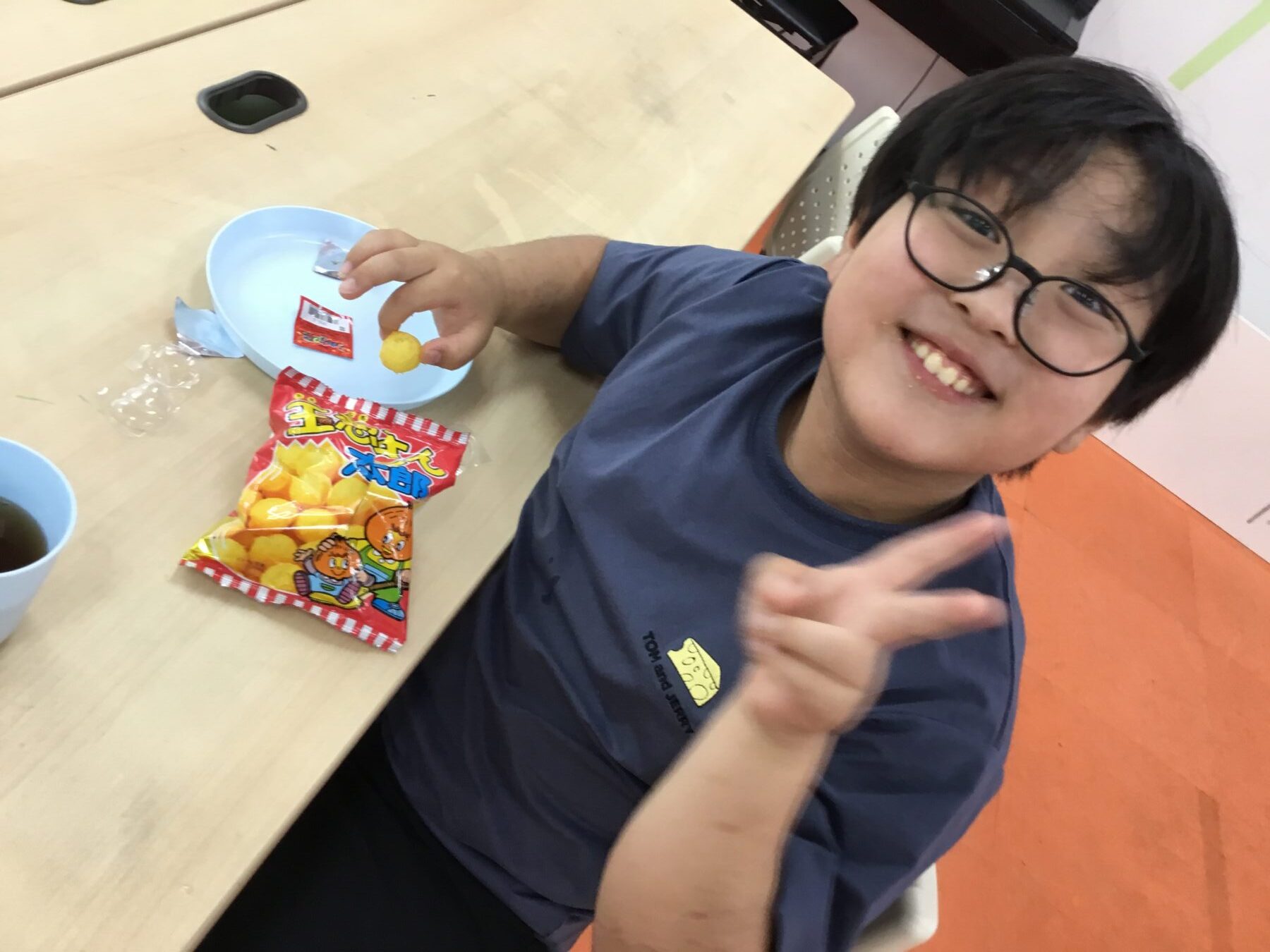 Child with glasses smiles at the camera while sitting at a table with a bag of snacks and a blue plate of yellow puff snacks nearby.