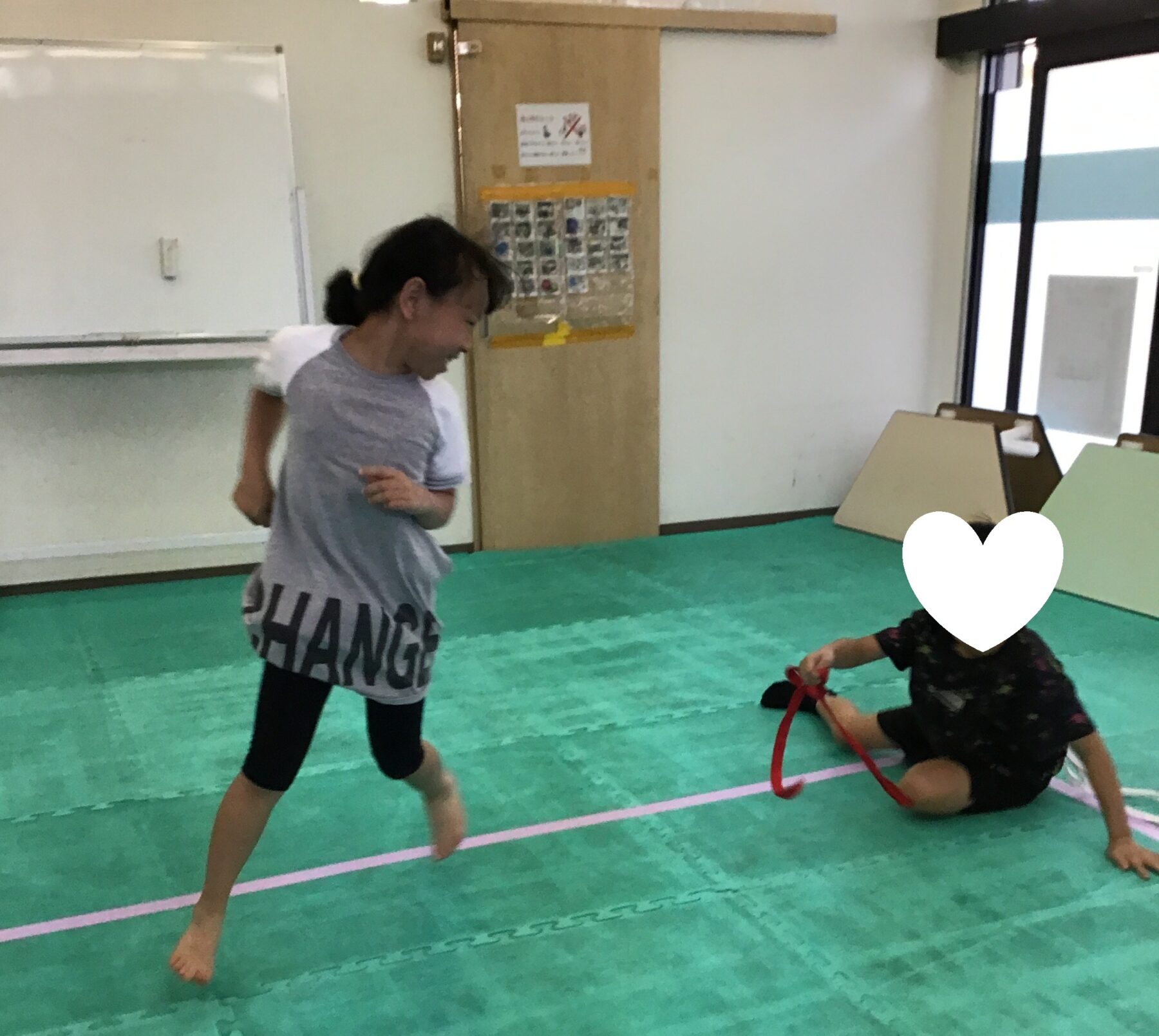 Two kids playing in a gym-like room on a green foam mat; one stands and runs while another sits on the floor with a red strap, a white heart graphic covers the seated child’s face.