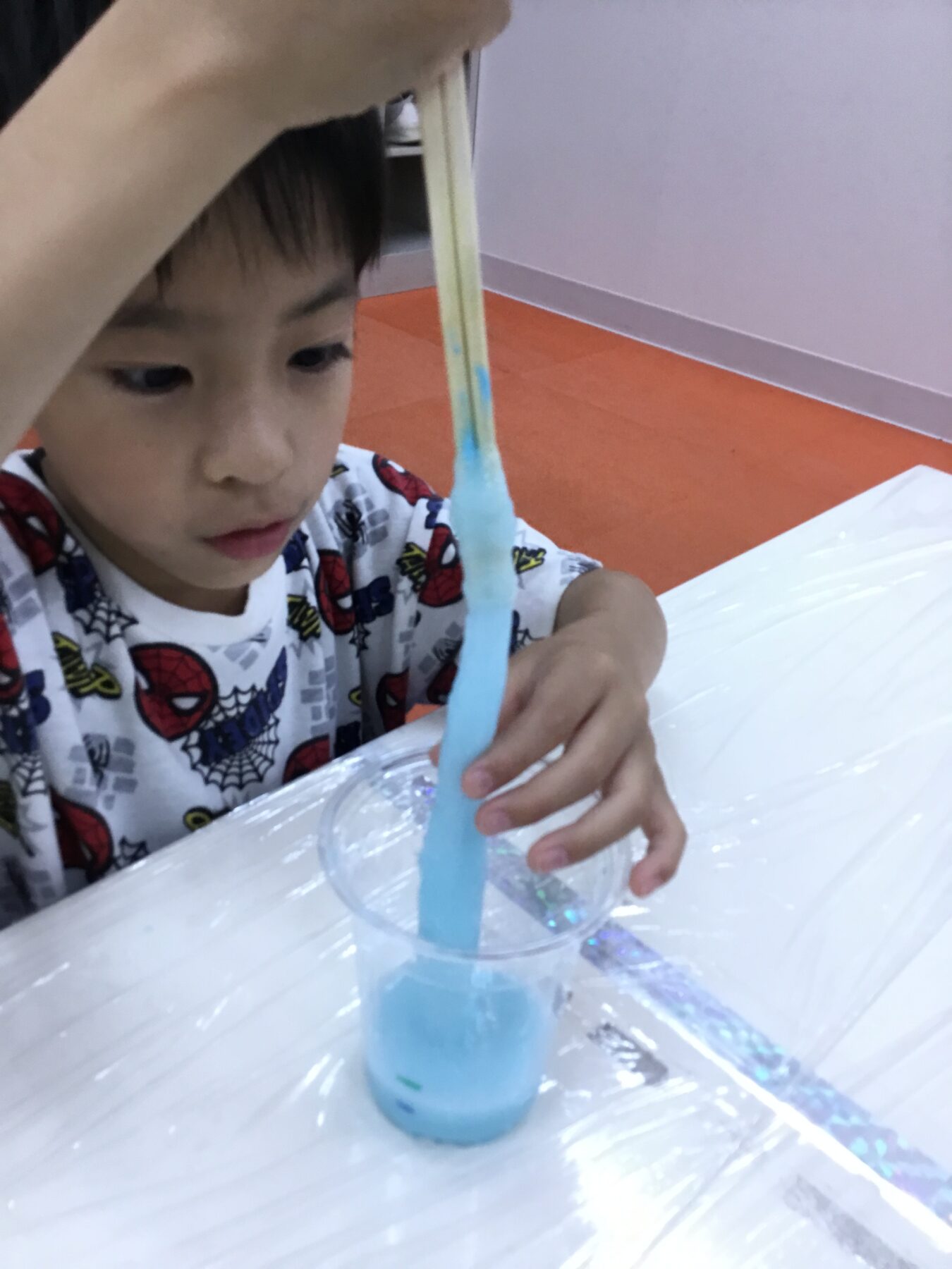 Young child in Spider-Man pajamas pours blue slime from a wooden stick into a clear cup on a white table.