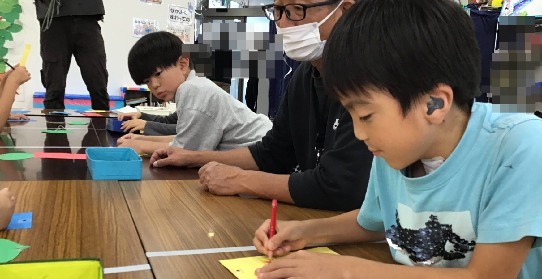 Children sit at a long table coloring with colored pencils, while an adult in a mask watches over the activity in a classroom.