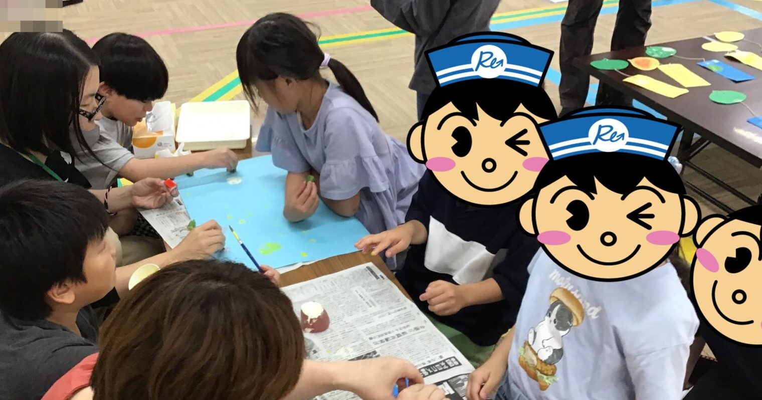 Group of children and adults seated around a table, painting on blue paper with brushes in a gym.