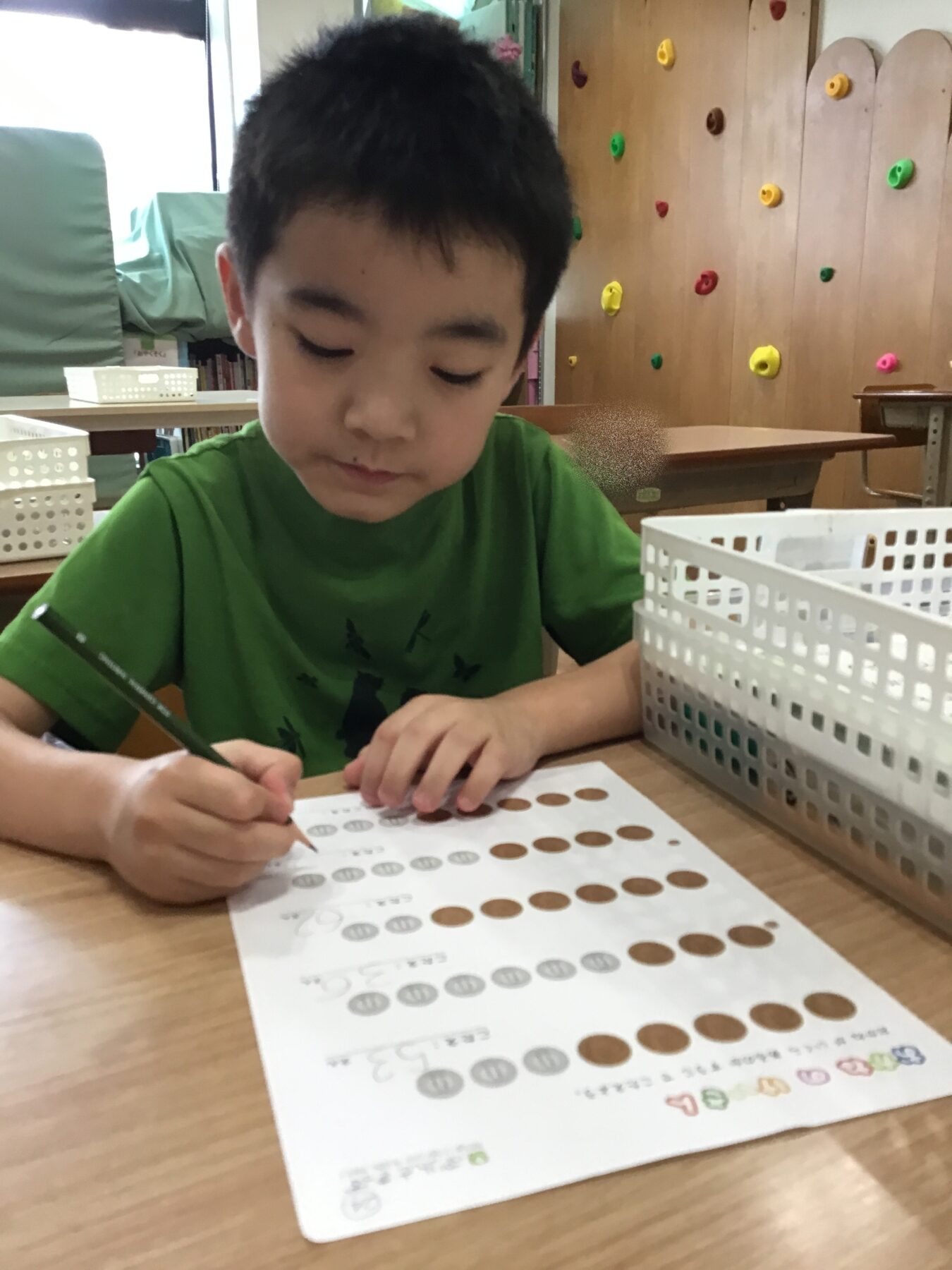 Young boy in a green shirt concentrates as he writes on a worksheet with dot patterns at a classroom table.