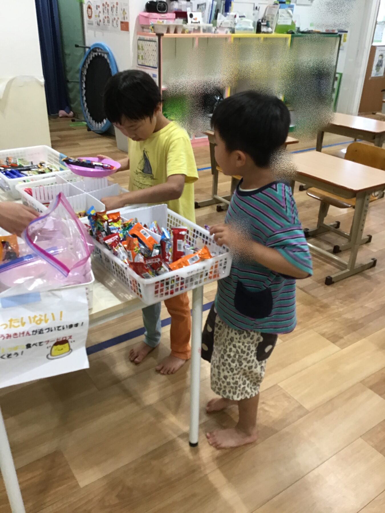 Two children in a classroom sorting colorful snack wrappers in white baskets on a table, one wearing a yellow shirt and the other striped shirt, both barefoot inside a bright room.