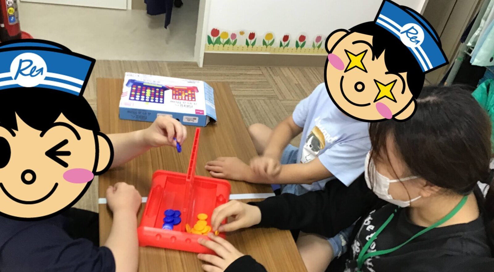 Children seated at a table playing a color-disk game with a red tray while an adult supervisor watches nearby in a classroom.