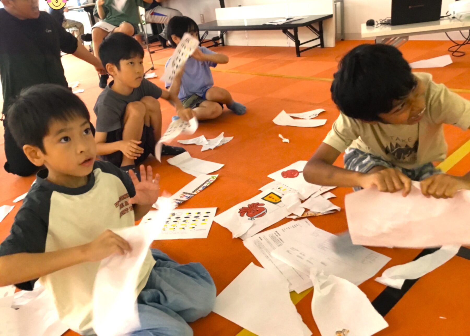 Children sitting on an orange gym floor doing paper crafts, with scraps and worksheets scattered around.