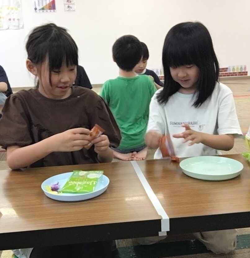 Two girls kneel at a low classroom table, opening snack packets as they share a treat. Bottles sit on the table; other kids in the background.