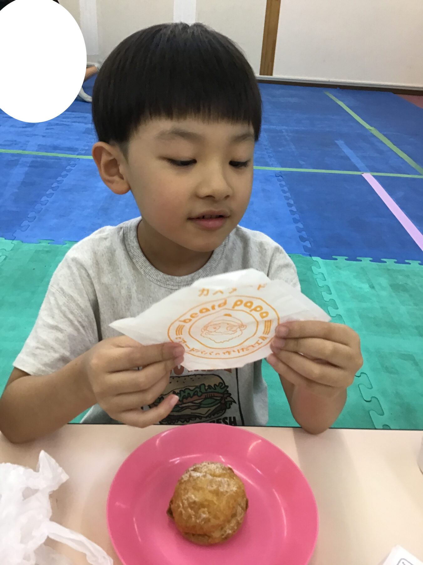Young boy with short black hair sits at a table, holding a white paper wrapper with an orange logo while a pink plate with pastry is in front of him on a colorful foam mat floor.
