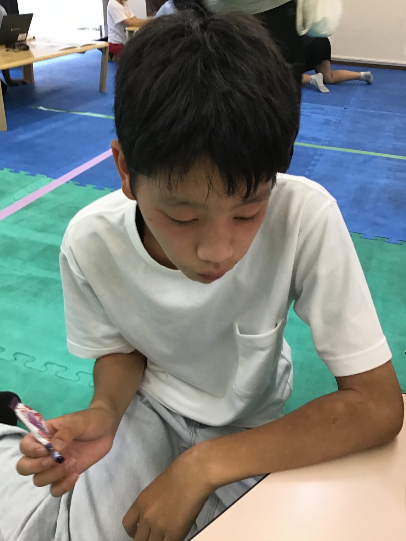 Boy in a white shirt sits on colorful foam mats, looking down at an object in his hand during an activity.