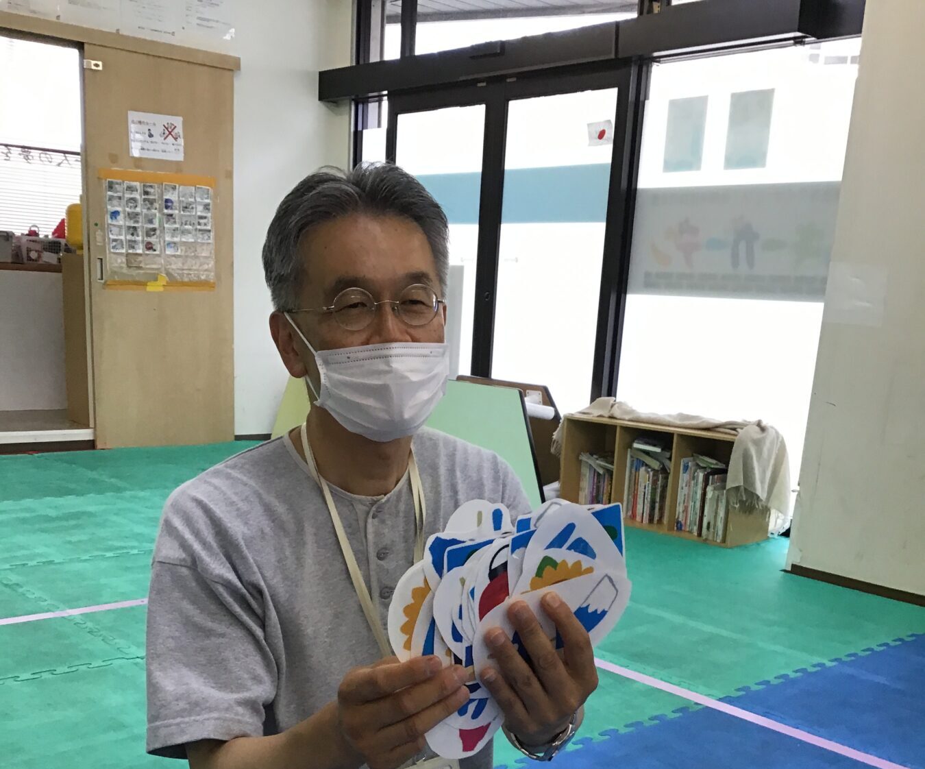 Man wearing a face mask and glasses kneels on colorful gym mats, holding a fan-shaped stack of bright paper cutouts.