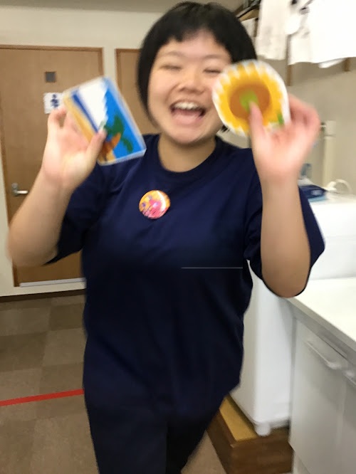 Smiling person with short black hair in a navy shirt, holding a blue card in one hand and a yellow circular plate in the other.