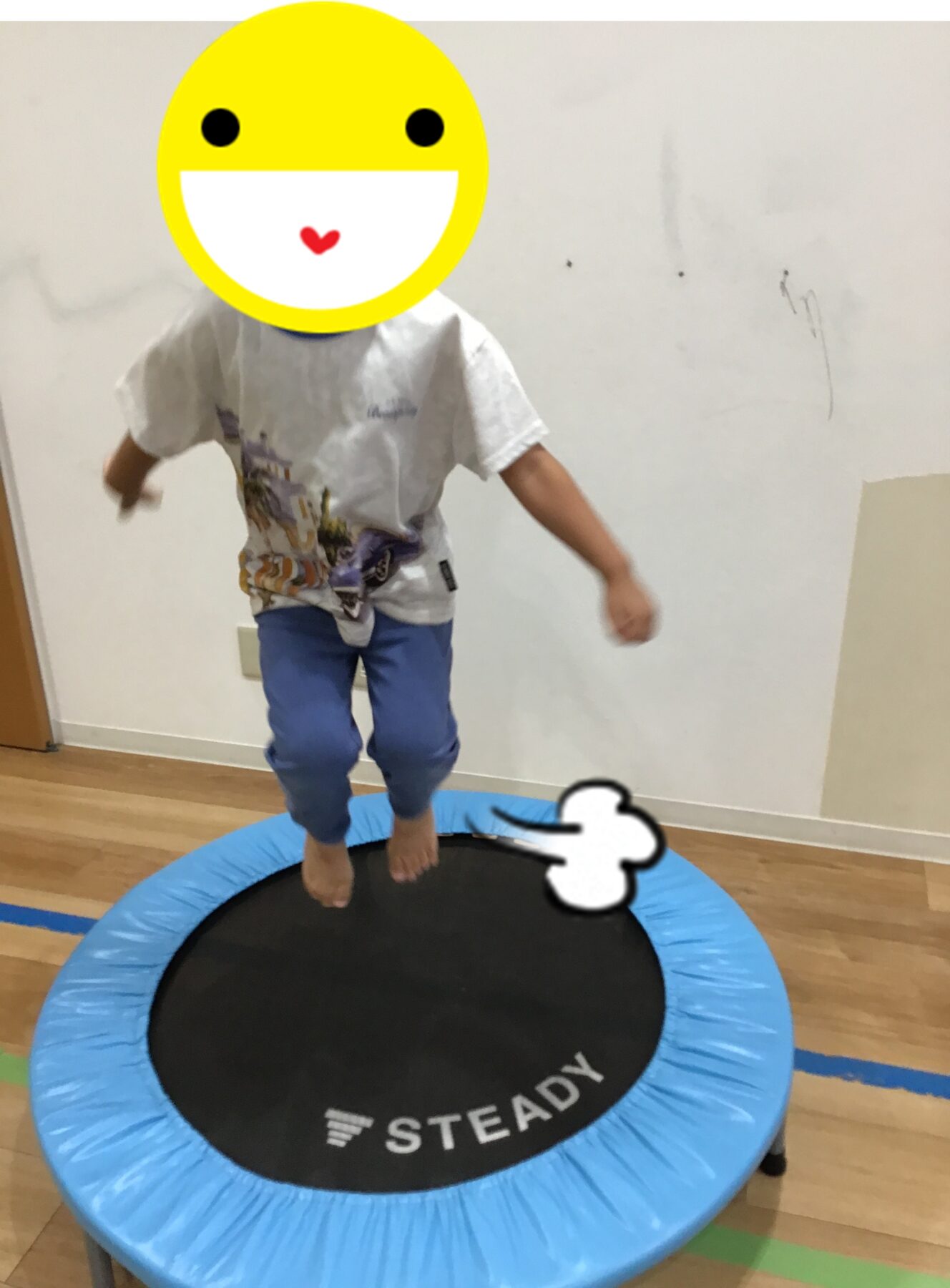 Child wearing a t‑shirt and blue pants jumps on a blue indoor trampoline; a yellow smiley overlay obscures their face; mat reads 'STEADY'.