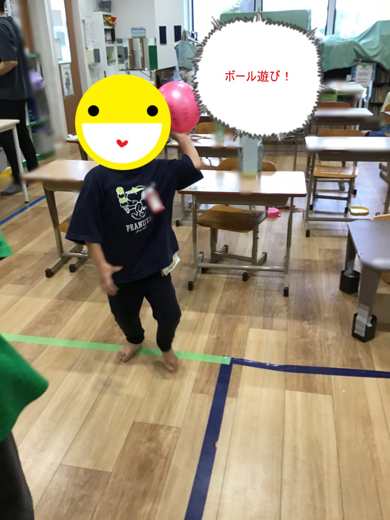 Child in a dark shirt holding a pink ball in a classroom-gym, with a large yellow emoji covering their face; speech bubble says ボール遊び! indicating ball play.